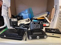 Front view of group of older model cameras including Kodak, Minolta, Nettar, and AGFAmatic, with Braun flash and camera cases