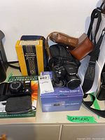 Lot overview showing Kodak Vigilant Junior Six-16 box, Canon PowerShot SX500 IS camera in box with camera on top, Minolta autofocus telephoto camera, photography book, and four empty camera cases spread on shelf.