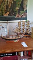 Two wooden model ships on a wooden surface with a landscape painting in the background. The smaller ship in front has white sails, the larger ship behind is labeled Flying Cloud with multiple beige sails.