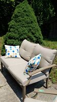 Outdoor metal-frame loveseat with beige cushions and two polka dot throw pillows, placed on concrete patio with a large green bush in the background.