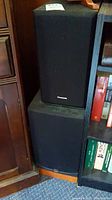 Two Panasonic speakers stacked vertically with black fabric grills, wooden base, and visible Panasonic logo on grills.
