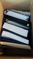 Photo of several large stamp albums stacked inside a box from a top view, showing thick pages mostly filled with stamps.
