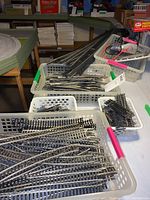 Photo of multiple white plastic baskets containing assorted model train track pieces, including straight, curved, and switch tracks, with shelving and a model train display partially visible in background.