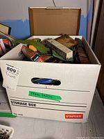 Open cardboard storage box with various train models, train tracks, and train-themed accessories packed inside.
