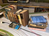 Wide shot showing a group of vintage hardback books arranged on a white surface against a basement wall. Books vary in size and color with visible titles and author names.