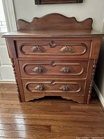 Front view of antique wash stand showing three drawers, two missing top drawer pulls, decorative carved wooden pulls on middle and bottom drawers, scalloped backsplash.
