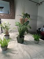 Four artificial plants of various types and sizes displayed on a surface with a mirror and side furniture in the background