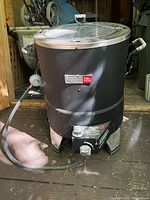 Angled view of the black cylindrical fryer with stainless steel edge, showing the propane control knob, handles, and branding.