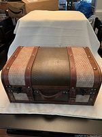 Front angle view of decorative storage trunk showing locking clasps, handle, and detailed textured material.