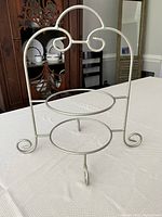 Photo of silver colored metal dessert plate stand on white textured surface, showing front view with two circular plate holders and decorative curved wire design.