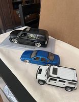 Three Dub City diecast cars on display: Cadillac Escalade (black), Chevy Monte Carlo (blue), and Hummer (white) on white cloth surface.