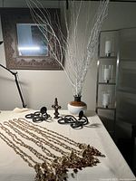 Full arrangement of all items on table including floor lamp base and arm, wall mirror reflecting a picture, metal sconces, decorative cross, ceramic vase with white sticks, and beaded tiebacks.