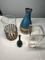 Four vases displayed on a white surface: a tall clear glass vase with circular metal decorations around it, a blue ceramic vase with a rope detail, a small dark textured metal vase, and a clear glass rectangular vase.