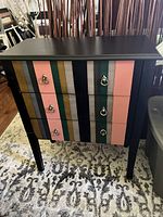Front view of the small black gloss three-drawer dresser with vertical multi-colored painted stripes and silver ring drawer pulls.