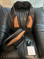 Black and brown cordless shoulder massager placed on black leather chair alongside charger and user manual. Soft padded texture with control buttons visible. Massager is supported on chair with its distinct shape for shoulders.