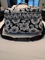 Front view of the Vera Bradley travel bag showing quilted black fabric with large gray and white Camellia flowers, two zippered compartments on the front and an adjustable black shoulder strap with padded section.
