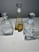 All three glass decanters shown together: amber colored, square clear with glass ball stopper, and square clear with unique stopper.