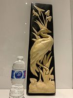 Full front view of the tall black plaque with large cream plaster bird figurine, shown with a water bottle for scale, illustrating the plaque's height and shape.