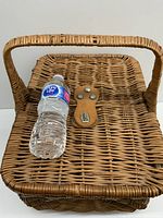 Front view of large vintage wicker basket with sturdy handle, showing hinged lid closed and secured by leather and metal lock plate. Pure Life water bottle placed on lid for scale.