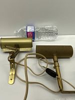 Two vintage brass cylindrical plug-in wall lamps with cords and inline switches laid side by side with a plastic water bottle for size reference.