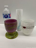Showcase of all three Venetian glass vessels grouped with a water bottle for size reference: purple rounded bowl with green base, tall white frosted vase, and clear bubbled textured bowl with swirl accent.