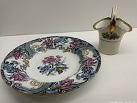 Photo showing both the antique floral plate with wear and the cream ceramic basket with floral decoration and gold trim.
