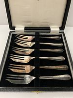 Six vintage Sheffield-plate forks displayed inside original black fitted case with velvet slots and white fabric lid lining.