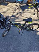 Photo of black children's bicycle with 16-inch wheels, neon yellow-green accents on saddle and frame. Bike is on pavement outside with shadows of tree branches and a metal chair nearby.