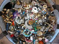 Overhead view of bag filled with mixed costume jewelry showing chains, beads, pendants, faux pearls, enamel charms and metal-tone pieces.