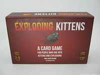Rectangular box of Exploding Kittens card game, brown with game title and cartoon cat print.
