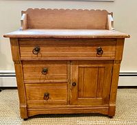 Front view of oak washstand showing scalloped backsplash, three drawers with bronze ring pull hardware, and single cabinet door with knob.