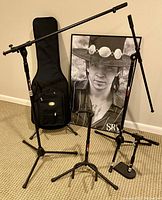 Wide shot of microphone stands, guitar case, guitar stand, amp stand and framed SRV portrait.