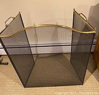 Black metal mesh fireplace screen with brass trimmed arched front and folding side panels with handles, standing on carpet.