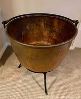 Large antique copper cauldron resting on a hammered cast iron tripod base, showing front and side profile, highlighting the patina and riveted handle detail.