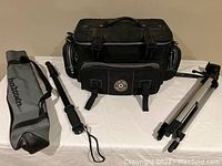 Black Kipling padded camera case with two small side pockets and a front pocket, accompanied by two tripods.
