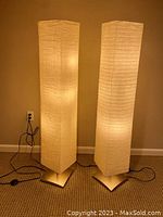 Two tall floor lamps side by side, lit, showing soft white light diffused by rectangular rice paper shades on brushed silver square domed bases.
