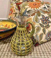 Photo of colorful hand-painted La Province Tuscan bowl, raffia-wrapped glass carafe with handle, and floral-patterned throw pillows on carpet.