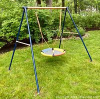 Front angle view of colorful metal swing set with blue legs and a colorful round swing disc suspended in the middle, on green grass with trees in background.