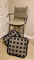Metal swivel bar stool with taupe cushioned back and seat with two black and gold patterned throw pillows in front.