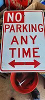 Photo shows a rectangular metal street sign measuring 14 by 10 inches with red text and border on a white background reading 'NO PARKING ANY TIME' with a double-headed red arrow at bottom.