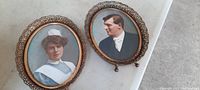 Front view of two antique oval framed photos featuring portraits of a nurse and a gentleman with bold coloration and glass dome fronts, placed on a light-colored surface.