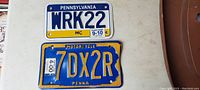 Photo showing two Pennsylvania motorcycle license plates: one older yellow and blue with embossed characters, and one modern white and blue with printed characters.
