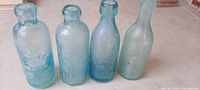 Four very old light blue glass bottles with embossed lettering and blob style tops arranged side by side.