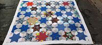 Full view of hand stitched quilt showing star hexagon pattern in blues, reds, and browns with white border on asphalt surface.