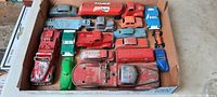 Top view of 15 antique steel toy vehicles in a cardboard box showing red Mobil tanker truck, red fire trucks, green and blue cars, several other trucks and cars all heavily used with paint wear.