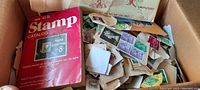 Photo of a boxed lot containing hundreds of loose mixed postage stamps with a red U.S. Stamp Catalog lying on top.