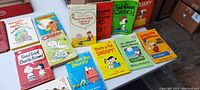 Front view of all comic character paperback books arranged on a surface showing covers with titles and illustrations of characters such as Snoopy, Dennis the Menace, and Charlie Brown.