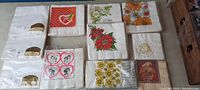 Twelve sealed vintage napkin packages laid out, showing front designs with holiday and floral themes such as hearts, flowers, and seasonal motifs.