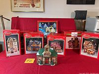 Full lot image showing six Dickens Collectables Towne Series lighted ceramic buildings with red boxes and one unboxed schoolhouse building.