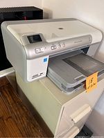 Front angle view of the HP Photosmart D5460 color inkjet printer on a filing cabinet, showing control panel, LCD screen, paper trays, and a sticky note labeled '18 HP Printer'.